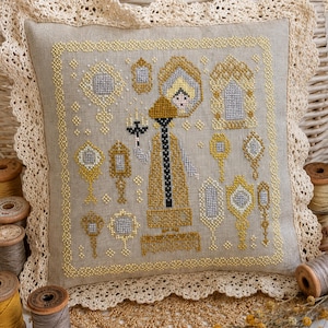 Puede incluir: Un coj&iacute;n cuadrado de lino beige con un dise&ntilde;o de punto de cruz que muestra una figura central sosteniendo un candelabro, rodeada de varios espejos de mano y marcos ornamentados en hilos dorados y plateados, con un borde de encaje crema.