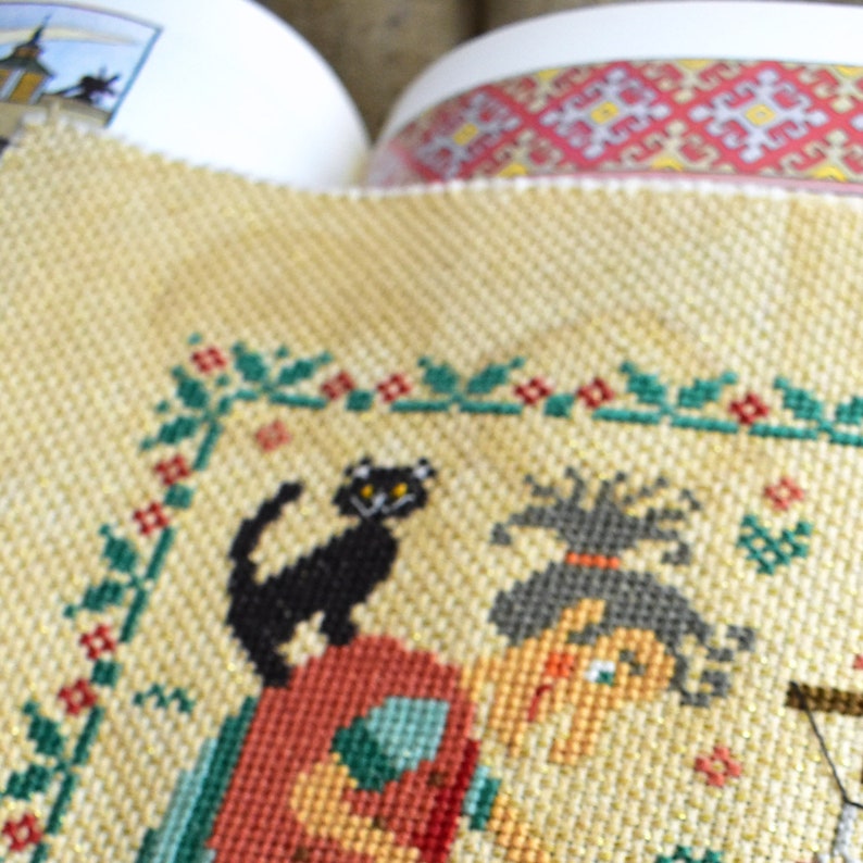 May include: A cross-stitch embroidery featuring a witch with a black cat and a green leafy border. The witch has a red and orange dress and a black hat. The background is a beige fabric.