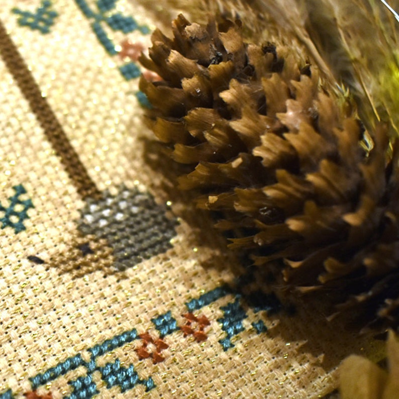 May include: A close-up of a brown pine cone on a beige fabric with a blue and gold cross-stitch pattern.