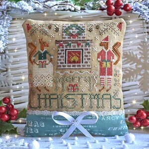 May include: A beige cushion with a cross-stitch design featuring two squirrels, a fireplace, and the text "Merry Christmas". The cushion is trimmed with white lace and tied with a white satin ribbon.