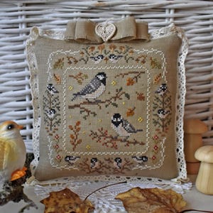 May include: A square beige fabric cushion featuring a cross-stitch design of birds on branches, surrounded by a cream-coloured lace border. A beige ribbon bow with a small crocheted heart sits at the top centre of the cushion.