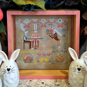 May include: A framed cross-stitch embroidery depicting two bunnies in a kitchen setting. The bunnies are wearing clothes and are preparing food. The embroidery is in a pink frame and is surrounded by two white bunny figurines.