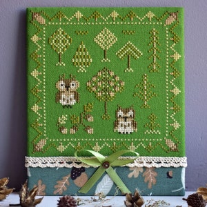 May include: A green cross-stitch embroidery featuring a woodland scene with owls, trees, and acorns. The embroidery is framed with a white lace trim and a green ribbon bow.