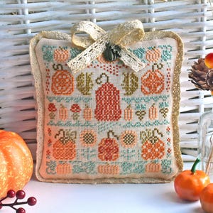 May include: A small, cream-colored pillow with a cross-stitch design featuring orange, red, and green pumpkins. The pillow has a lace trim and a brown ribbon tied around it.