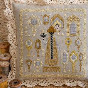 Puede incluir: Un coj&iacute;n cuadrado de lino con un dise&ntilde;o de punto de cruz en tonos dorados, amarillos y grises. El bordado muestra una figura central sosteniendo un candelabro rodeada de varios espejos de mano y marcos ornamentados, con borde de encaje.