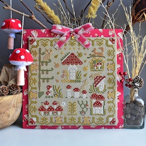 Peut inclure: Une broderie au point de croix représentant une scène de forêt fantaisiste avec des champignons, des écureuils et une petite maison. La broderie est encadrée et a une bordure à carreaux rouge et blanc.