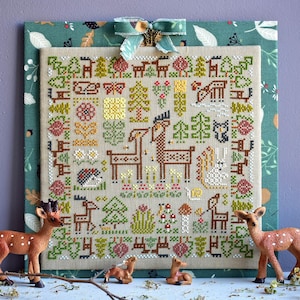 May include: A cross-stitch pattern depicting a woodland scene with deer, trees, and flowers. The pattern is stitched on a beige fabric with a green border. The pattern is surrounded by small deer figurines.