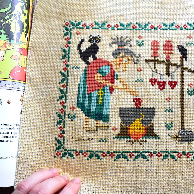 May include: Cross-stitch embroidery of a witch stirring a cauldron with a black cat on her shoulder. The witch is wearing a red and blue dress and a white apron. The cauldron is over a fire and there are three red apples hanging on a stick in the background.