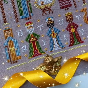 K&ouml;nnte beinhalten: Ein Kreuzstichmuster mit den Heiligen Drei K&ouml;nigen, mit detaillierten Stichen in verschiedenen Farben. Das Design enth&auml;lt das Wort "NATIVITY" und wird durch eine goldene Engelsfigur und ein gelbes Band akzentuiert.