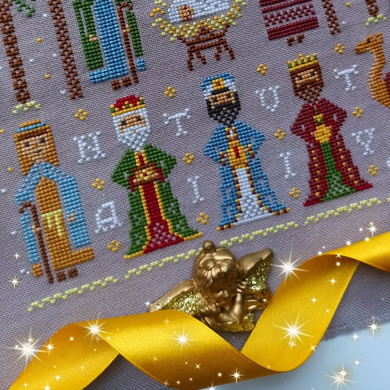 K&ouml;nnte beinhalten: Ein Kreuzstichmuster mit den Heiligen Drei K&ouml;nigen, mit detaillierten Stichen in verschiedenen Farben. Das Design enth&auml;lt das Wort "NATIVITY" und wird durch eine goldene Engelsfigur und ein gelbes Band akzentuiert.