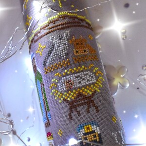 K&ouml;nnte beinhalten: Ein kreuzgestickter Weihnachtsschmuck mit einer Krippenszene. Das Design umfasst einen Stall, Tiere und das Jesuskind, alles in verschiedenen Farben auf einem grauen Stoff. Der Schmuck ist zylindrisch und mit gelben Sternen verziert.