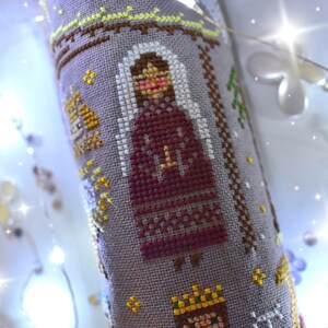 K&ouml;nnte beinhalten: Ein Kreuzstich-Design auf grauem Stoff, das eine Figur in einem bordeauxfarbenen Kleid und wei&szlig;em Schleier zeigt. Das Design enth&auml;lt gelbe und braune Akzente mit einem Kreuz auf dem Kleid der Figur. Das Gesamtthema ist religi&ouml;s.