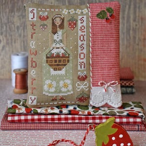 May include: A rectangular fabric pincushion featuring a cross-stitch design of a person holding a basket of strawberries, with the text "strawberry season" embroidered vertically. The side panel is red and white gingham with a white lace trim and bow.