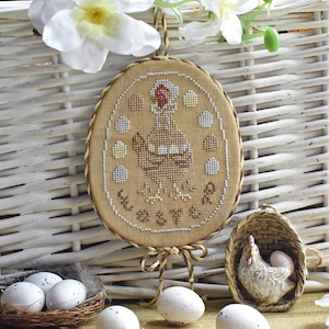 May include: A brown and white embroidered oval ornament with a brown hen and eggs, surrounded by a brown rope border. The ornament reads "Happy Easter".