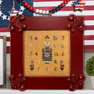 May include: A framed cross-stitch sampler with a patriotic theme. The red frame surrounds a tan fabric with a stitched design of a person, flags, and the word "red". Decorative elements include a star and a beaded garland.