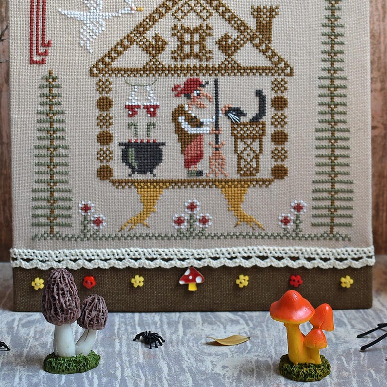 May include: A cross-stitch sampler depicts a witch inside a house, with a black cat, cauldron, and broom. The sampler is framed with a brown border and decorated with mushroom and flower figurines. The color palette includes beige, brown, red, and black.