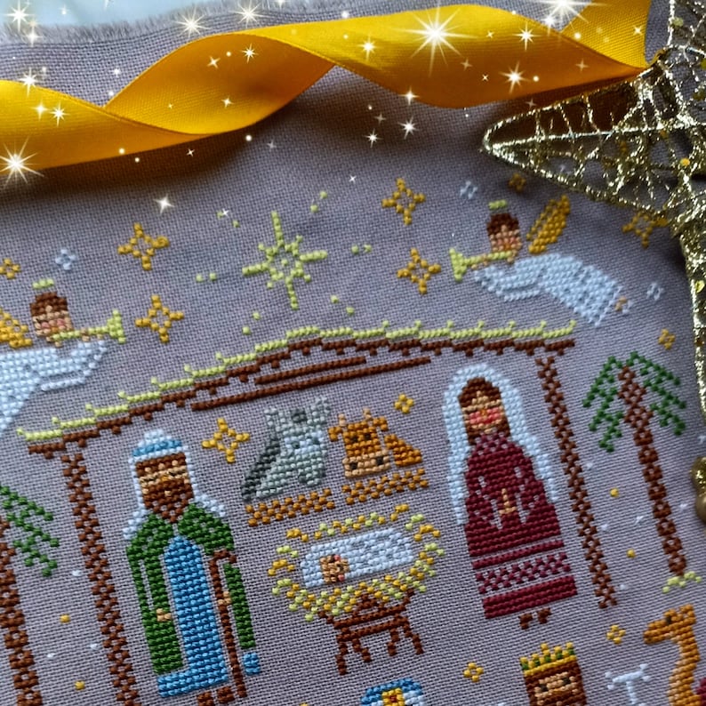 K&ouml;nnte beinhalten: Kreuzstich-Stickerei, die eine Weihnachtskrippe mit Engeln, der Heiligen Familie und den Heiligen Drei K&ouml;nigen darstellt. Das Design weist eine ged&auml;mpfte Farbpalette mit Gold- und Gelb-Akzenten und ein goldenes Band auf.