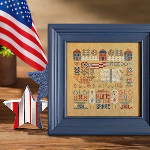 May include: A framed cross-stitch sampler with a patriotic theme. The sampler features houses, an American flag, and the words "Freedom", "Old", "Home of the Brave", and "Glo". A blue star and a star with red and white stripes are in the foreground.