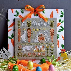 May include: A cross-stitch embroidery design featuring a bunny rabbit, carrots, and eggs. The design is framed and displayed on a white background with a carrot pattern.