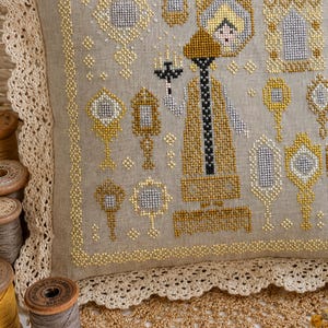 Puede incluir: Un coj&iacute;n de lino beige con un patr&oacute;n de punto de cruz que muestra una figura central sosteniendo un candelabro, rodeada de varios espejos de mano ornamentados en hilos dorados y plateados, con un borde de encaje de color crema.