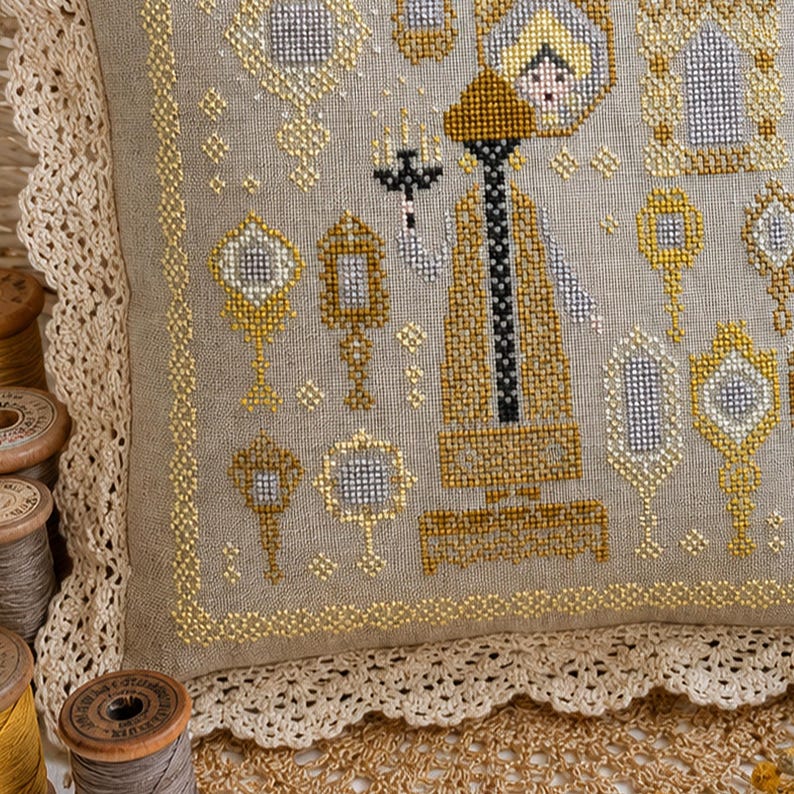 Puede incluir: Un coj&iacute;n de lino beige con un patr&oacute;n de punto de cruz que muestra una figura central sosteniendo un candelabro, rodeada de varios espejos de mano ornamentados en hilos dorados y plateados, con un borde de encaje de color crema.