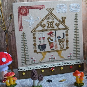 May include: Cross-stitch embroidery featuring a whimsical witch's house scene. The design includes a witch, a black cat, a cauldron, and a goose. The color palette is dominated by beige, brown, red, and white, with decorative elements.