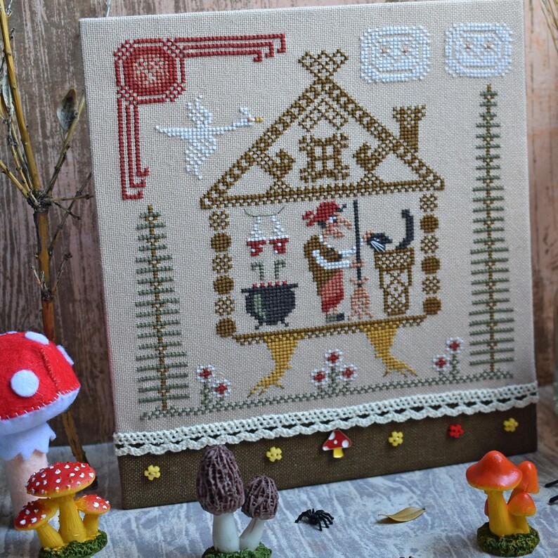 May include: Cross-stitch embroidery featuring a whimsical witch's house scene. The design includes a witch, a black cat, a cauldron, and a goose. The color palette is dominated by beige, brown, red, and white, with decorative elements.
