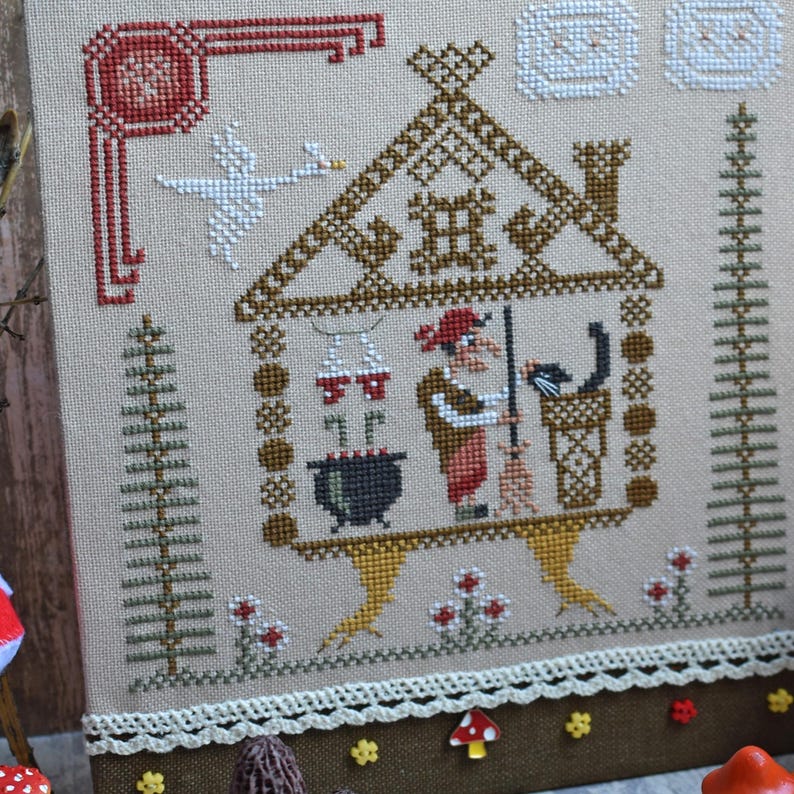 May include: Cross-stitch sampler featuring a whimsical scene with a house, a witch, and a black cat. The design includes a cauldron, a swan, and decorative borders. The color palette includes brown, red, and white on a beige background.
