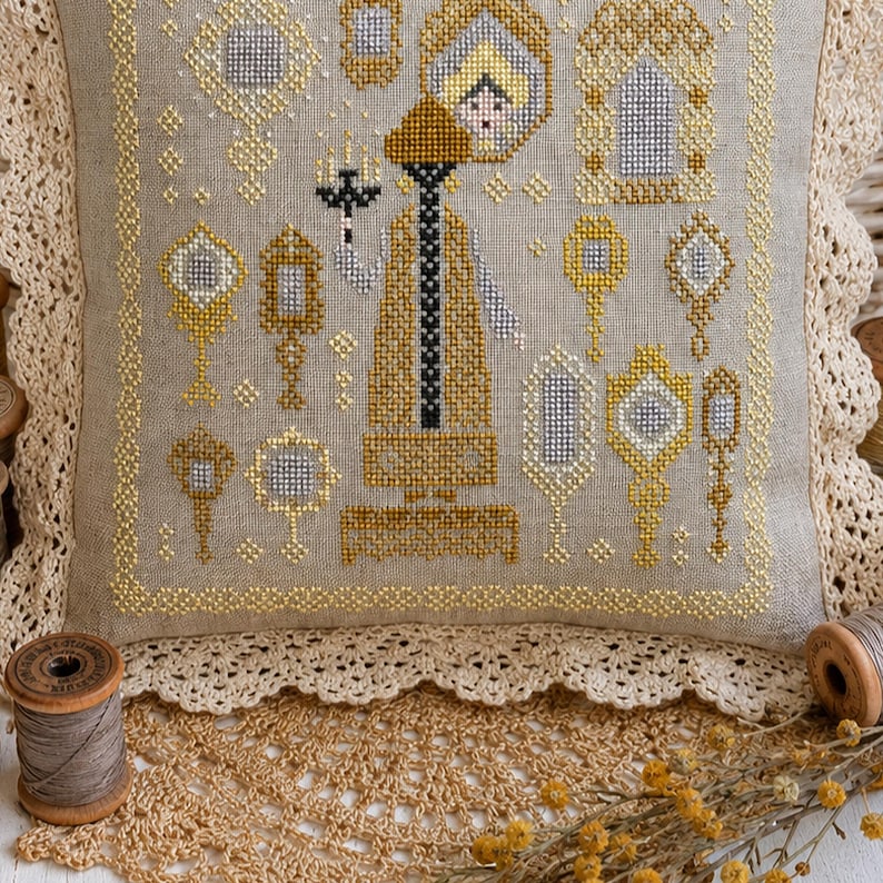 Puede incluir: Un coj&iacute;n cuadrado de lino con un dise&ntilde;o de punto de cruz que muestra una figura central sosteniendo un candelabro, rodeada de varios espejos de mano ornamentados en tonos dorados, plateados y negros, con un borde de encaje color crema.