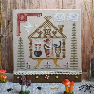 May include: Cross-stitch embroidery featuring a witch in a house with a black cat, cauldron, and broom. The artwork has a beige background with brown and red accents, and a white lace trim. Decorative mushrooms and spiders are in the foreground.