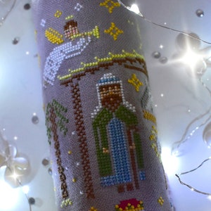 K&ouml;nnte beinhalten: Ein Kreuzstichstoff mit einer Weihnachtskrippe. Das Design umfasst einen Engel, der Trompete spielt, einen Mann in einer Robe, Palmen und Sterne. Die Farbpalette besteht aus grauen, wei&szlig;en, gelben, gr&uuml;nen und braunen F&auml;den und erzeugt ein detailliertes, festliches Bild.