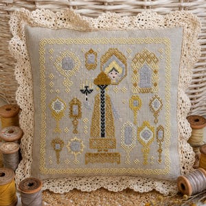 Puede incluir: Un coj&iacute;n cuadrado de lino beige con un dise&ntilde;o en punto de cruz de una figura central que sostiene un candelabro, rodeada de varios espejos de mano ornamentados en tonos dorados, plateados y negros, con un borde de encaje de ganchillo crema.