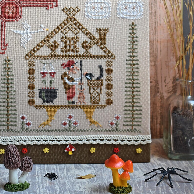 May include: A cross-stitch sampler featuring a witch in a house, with a black cat, cauldron, and goose. Decorative mushrooms, a spider, and other embellishments are in the foreground. The color palette includes brown, beige, red, and white.