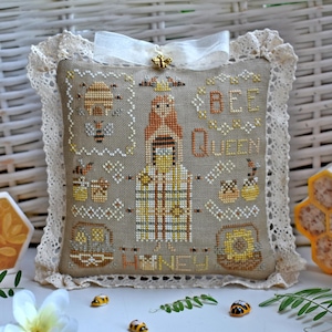 May include: A decorative pillow with a cross-stitch design featuring a bee queen theme. The pillow has a beige background with embroidered bees, honeycombs, and the words "BEE QUEEN" and "HONEY". It is trimmed with lace and a white ribbon.