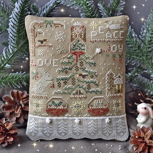 May include: A beige cross-stitch pillow with a Christmas tree, baskets, and the words "LOVE", "PEACE", and "JOY". The pillow has a white lace trim and is decorated with a star and a small house.