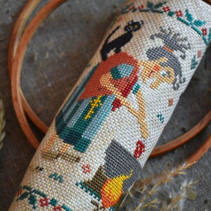 May include: A cross-stitch embroidery of a witch with a black cat, a cauldron, and a pumpkin. The witch is wearing a red and blue dress and has a black hat. The background is a cream-colored fabric with a gold glitter finish.