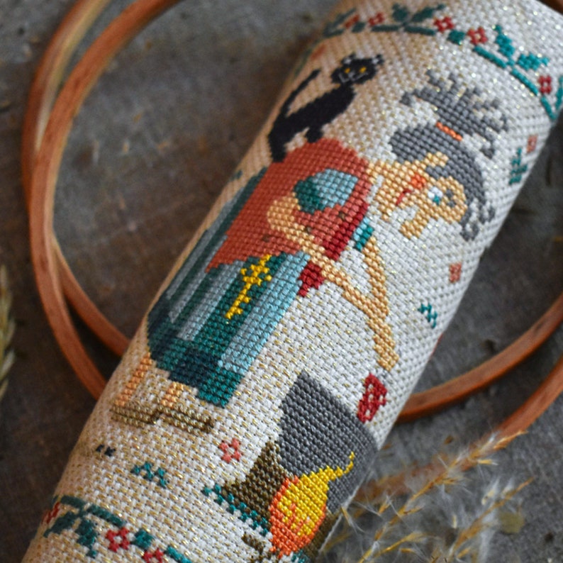 May include: A cross-stitch embroidery of a witch with a black cat, a cauldron, and a pumpkin. The witch is wearing a red and blue dress and has a black hat. The background is a cream-colored fabric with a gold glitter finish.