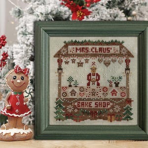 May include: A framed cross-stitch depicting Mrs. Claus's Bake Shop, with the text "MRS. CLAUS" and "BAKE SHOP". The artwork is in a green frame, and a gingerbread woman figurine is in the foreground. The background includes a snowy Christmas tree.