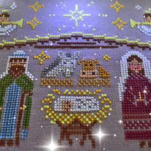 K&ouml;nnte beinhalten: Kreuzstich-Stickerei, die eine Weihnachtskrippe darstellt. Das Kunstwerk zeigt die Figuren von Josef, Maria und dem Jesuskind in einer Krippe, zusammen mit einem Ochsen, einem Esel und Engeln. Die Farbpalette umfasst Braunt&ouml;ne, Gr&uuml;n, Gelb und Rot.