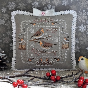 May include: A gray, square decorative pillow with a cross-stitched design featuring two birds perched on a branch. The pillow has a white lace trim and a white bow with a pink button. The background has a snowflake pattern.