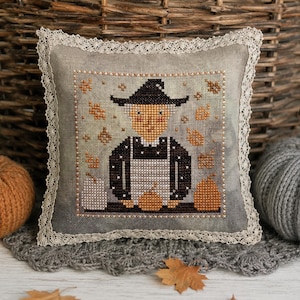 May include: A square decorative pillow featuring a cross-stitch design of a person wearing a black hat and apron, surrounded by orange autumn leaves and pumpkins. The pillow is edged with cream-colored lace trim on all four sides.