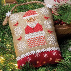 May include: A Christmas-themed, cross-stitched ornament featuring Santa Claus in a chimney, with red cardinals and snowflakes. The ornament has a burlap-like background, a red bottom border, and a rope handle.