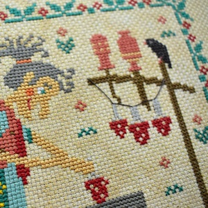 May include: Cross-stitch embroidery featuring a woman with brown hair and a red dress, holding a red cup. The background includes a brown wooden post with red birds and a black bird.