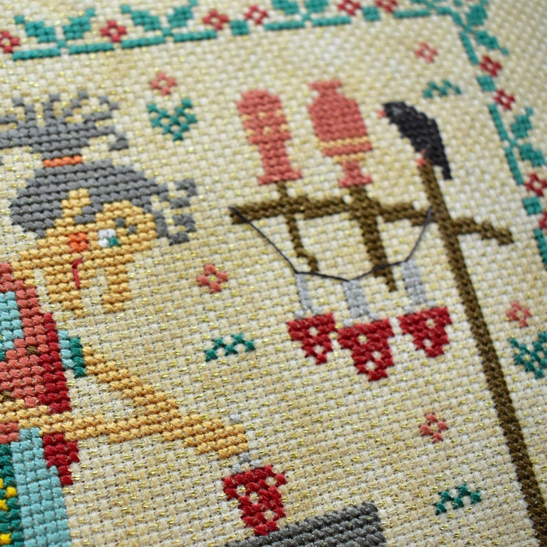 May include: Cross-stitch embroidery featuring a woman with brown hair and a red dress, holding a red cup. The background includes a brown wooden post with red birds and a black bird.
