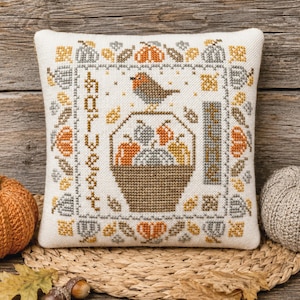 May include: A square off-white fabric pillow featuring a cross-stitch design of a brown bird perched on a basket filled with orange, yellow, and gray pumpkins. The text HARVEST TIME is stitched vertically in brown, surrounded by a border of leaves.