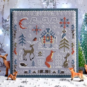 May include: A framed cross-stitch depicting a winter landscape with a grey backdrop. The design includes a crescent moon, snowflakes, a house, trees, and animals such as a deer, fox, and rabbit. The frame is blue.
