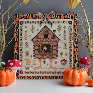 May include: A cross-stitch pattern of a witch's house on chicken legs. The house is brown with a red roof and a black cat in the window. The house is surrounded by green trees and flowers. The pattern is framed in a black and orange fabric with a mushroom pattern.