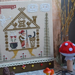 May include: Cross-stitch embroidery featuring a witch in a house with a cauldron, broom, and cat. The artwork is in shades of brown, beige, red, and white. A decorative border and small mushroom accents are at the bottom. A red mushroom decoration is in the foreground.