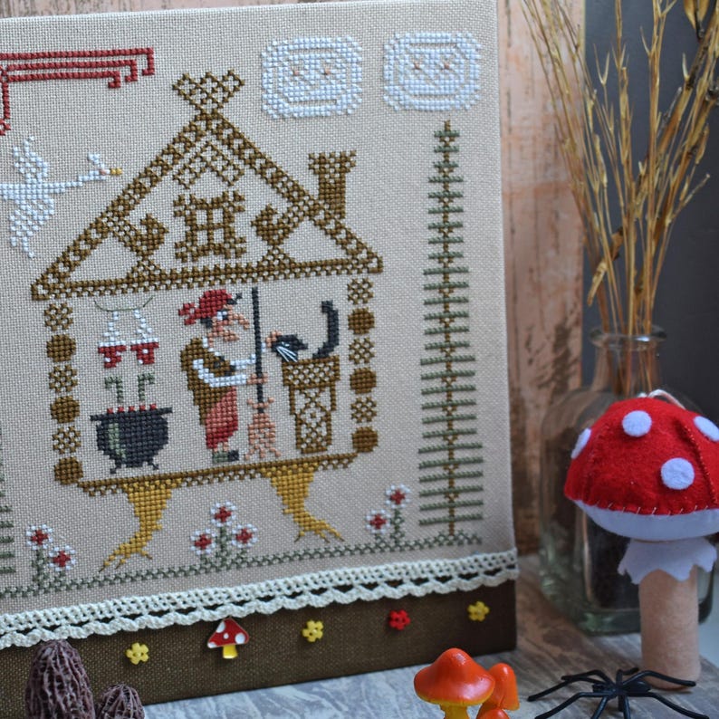 May include: Cross-stitch embroidery featuring a witch in a house with a cauldron, broom, and cat. The artwork is in shades of brown, beige, red, and white. A decorative border and small mushroom accents are at the bottom. A red mushroom decoration is in the foreground.