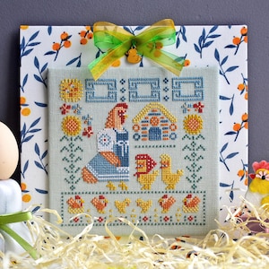 May include: Cross-stitch embroidery of a hen and chicks in a green and blue farmhouse setting. The hen is wearing a blue dress and holding a red flower. The chicks are yellow and have black eyes. The background is a light green with a blue farmhouse and a red flower.